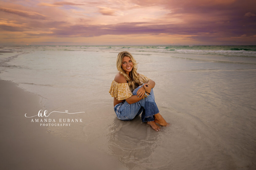 A smiling senior girl sitting on the shoreline with pastel skies overhead during a Anna Maria Island senior photography session