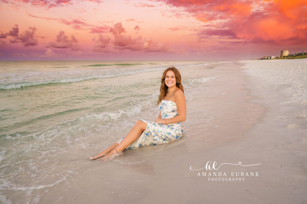 A senior girl in a white dress seated at the shoreline beneath pink skies, photographed by a Fort De Soto senior photographer