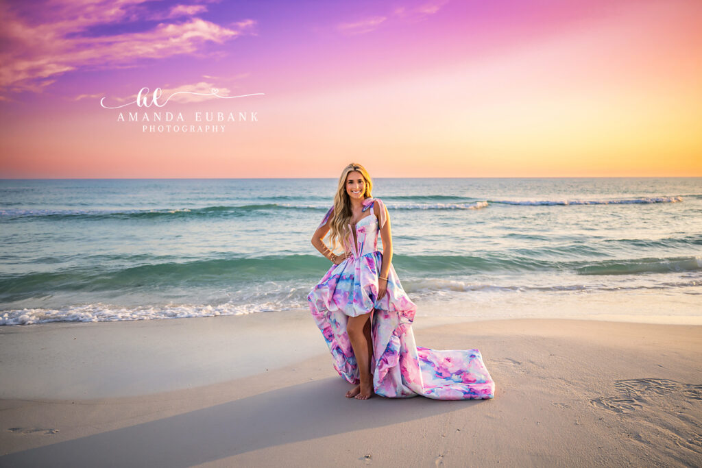 A woman in a flowing floral gown standing at the shoreline beneath a purple sky during a Redington Beach portrait photography session