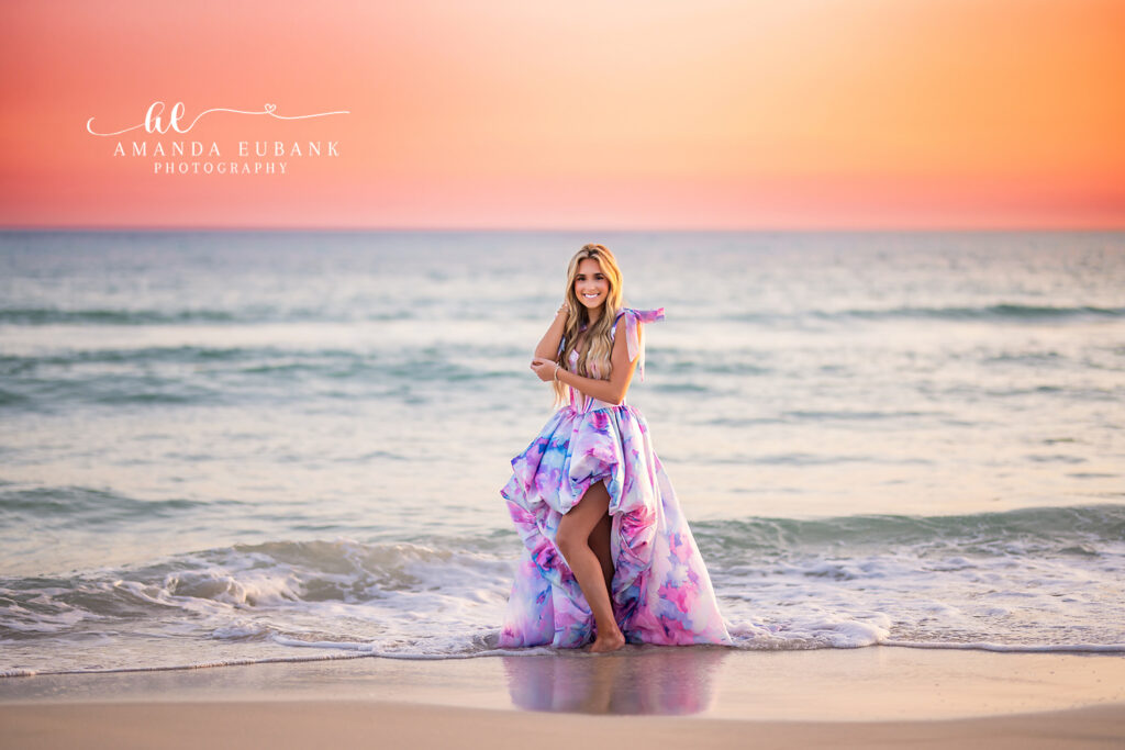 A senior girl in a lavender high-low dress standing at the shoreline in pastel light in Sarasota, Florida