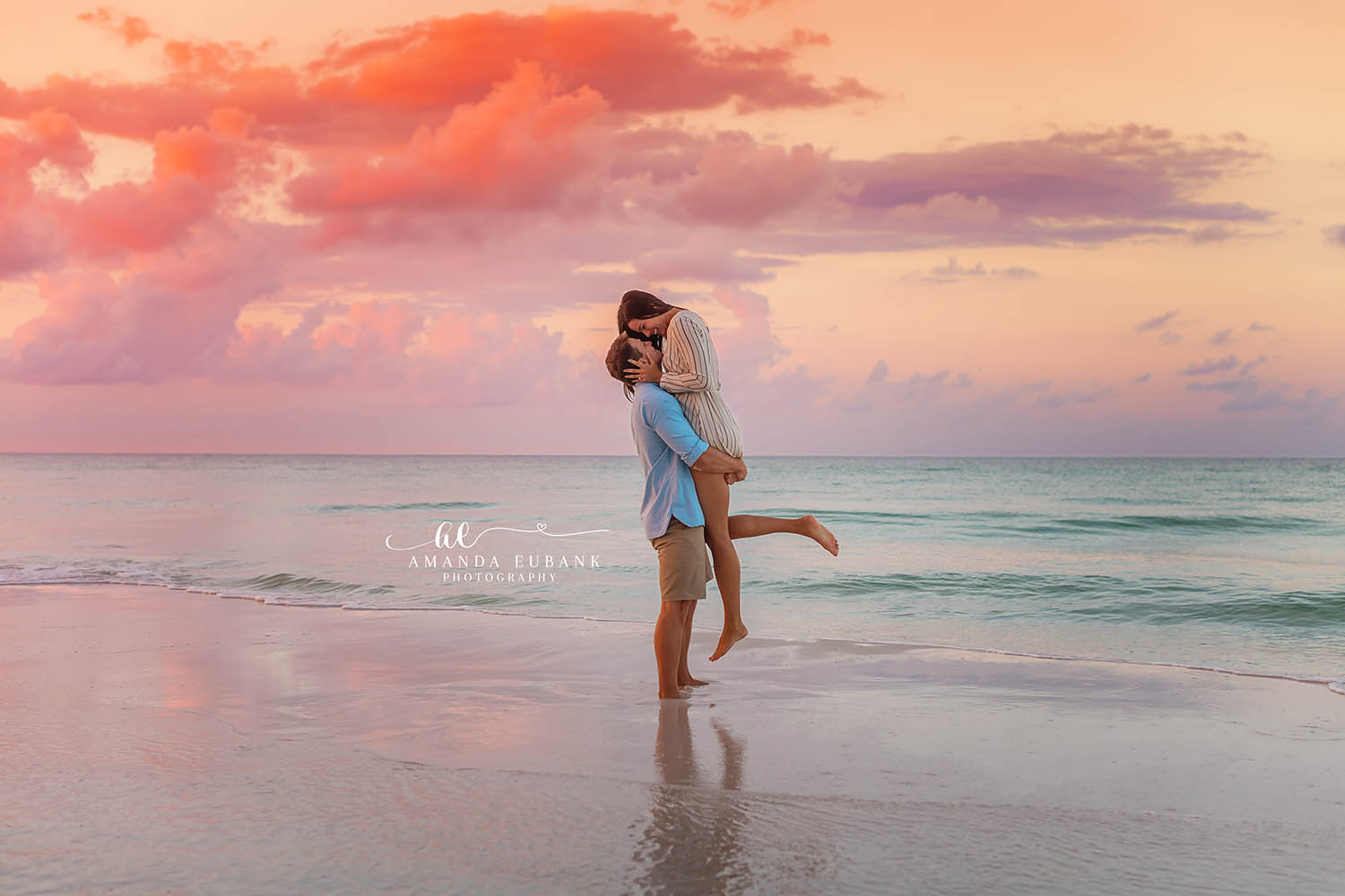 Sunset Lift on the Shoreline — Tampa, FL Engagement Session A man lifting his fiancee off her feet at the water's edge under pink sunset clouds at a Gulf beach engagement session near Tampa, Florida.