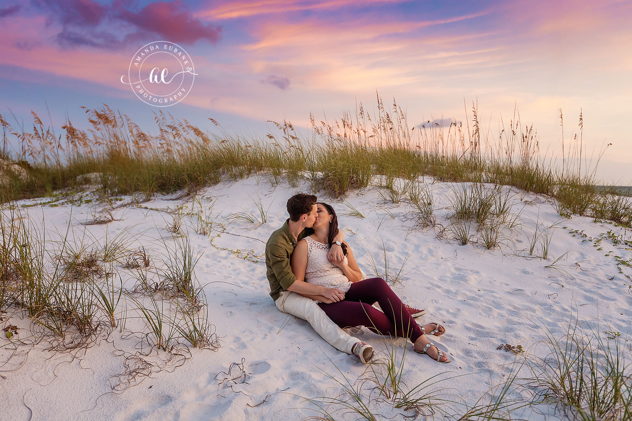 Golden Hour in the Sand Dunes — Tampa, FL Engagement Session A couple kissing while reclining on a white sand dune fringed with sea oats at a Gulf beach engagement session near Tampa, Florida.
