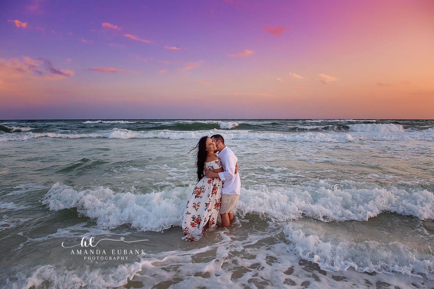 Kissing in the Breaking Waves — Tampa, FL Engagement Session A couple kissing while waves break around them under a pink and purple sky at a Gulf beach engagement session near Tampa, Florida.