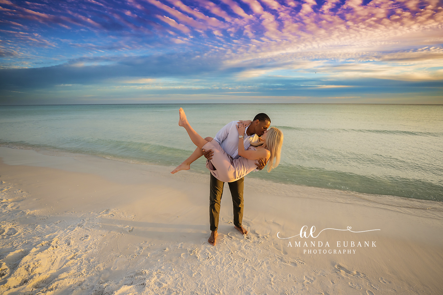 Carried Across the Sand | Engagement Photographer in Tampa, Florida Engagement photos in Tampa, FL showing a man carrying his fiancee across white sand beneath a rippled blue cloudscape.