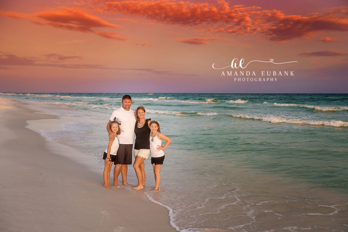 Standing in the Surf — Tampa, FL Family Session A family of four standing together in the shallow surf under a coral sky at a beach family session near Tampa, Florida.