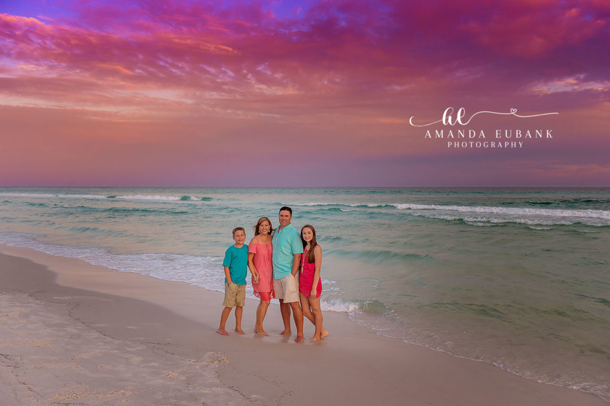 Family at the Water’s Edge — Tampa Bay Family Portraits A family of four in teal and coral standing at the water's edge at dusk — beach family photography in Tampa Bay.