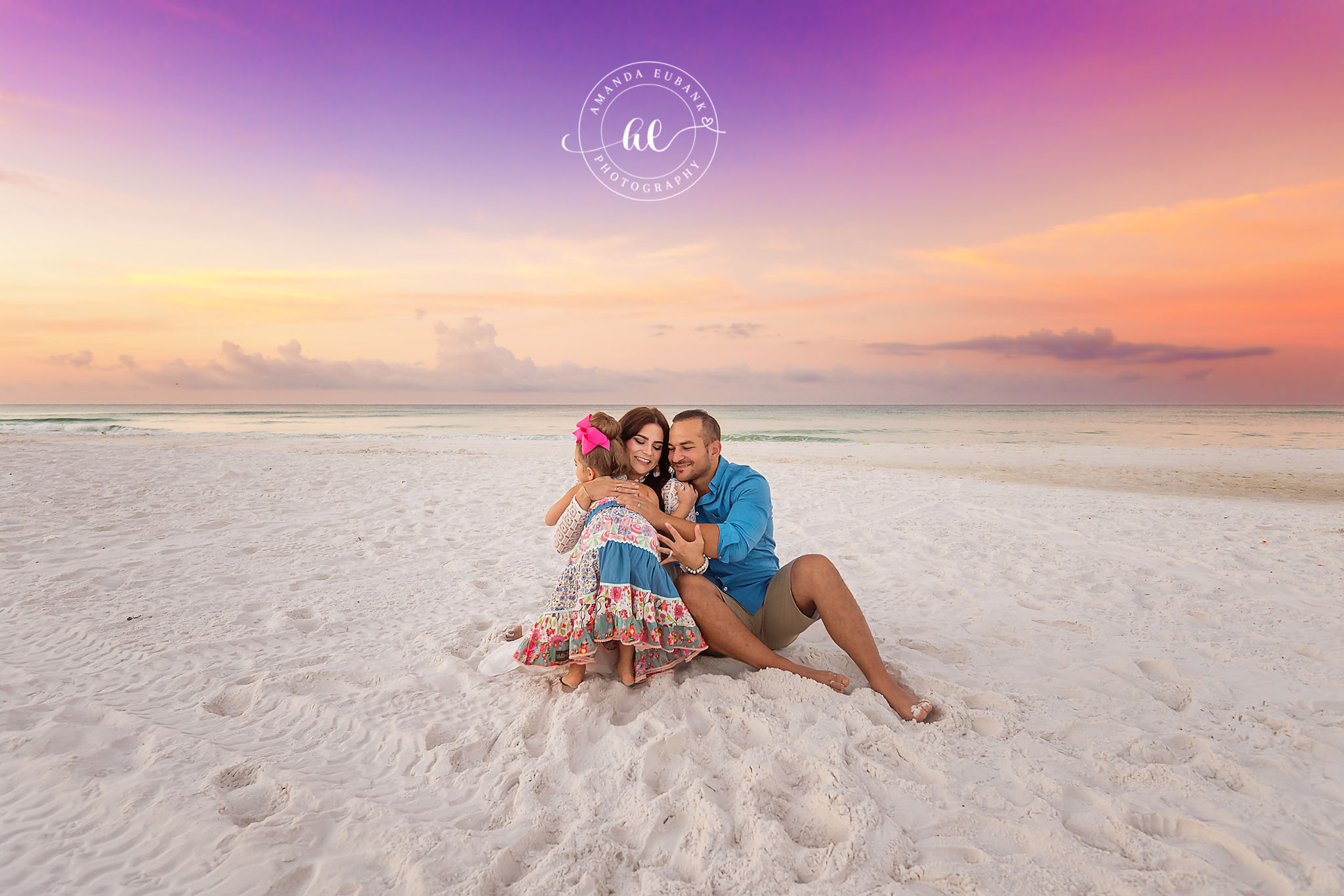 Family with a Toddler | Tampa Beach Family Photography Tampa family photographer captures parents and their toddler sitting on the sand beneath a purple and orange sky.