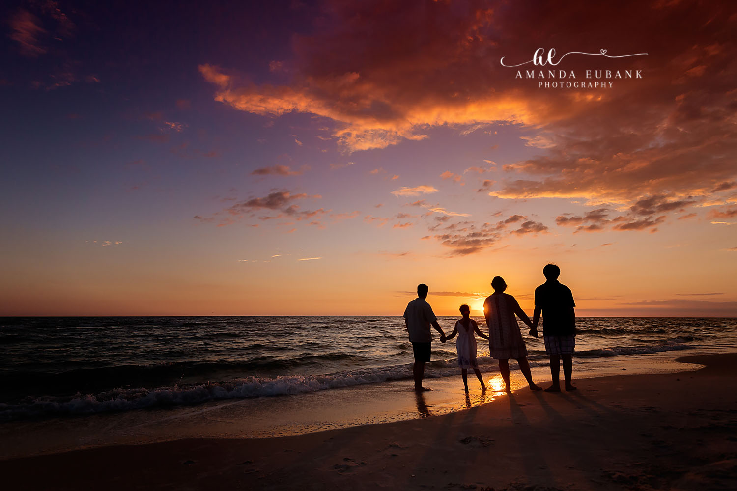 Family Silhouette at Sundown | Family Photographer in Tampa, Florida Family beach portraits in Tampa, FL showing a family of four silhouetted holding hands as the sun touches the horizon.