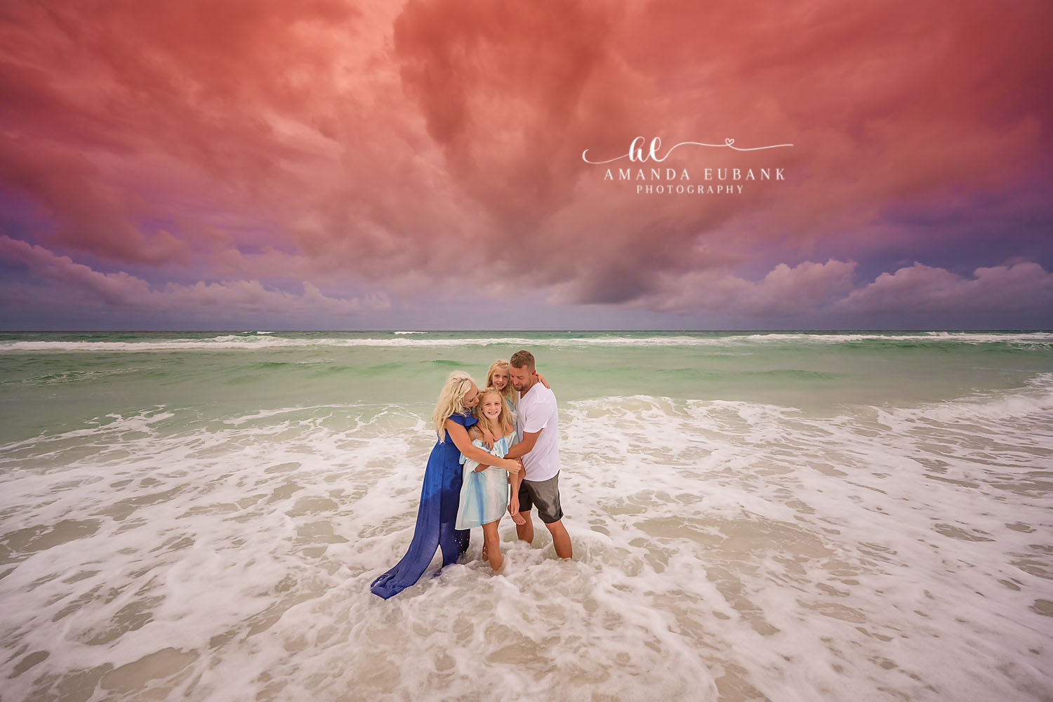 Sitting in the Shallow Surf | Tampa Beach Family Photography Tampa family photographer captures a family of four sitting in the shallow surf beneath dramatic pink clouds.
