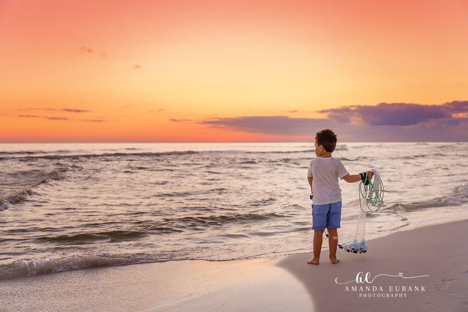 Boy With a Net | Kids Beach Portraits in Tampa, FL Tampa children’s photographer captures a young boy standing at the shoreline holding a net under an orange sky.