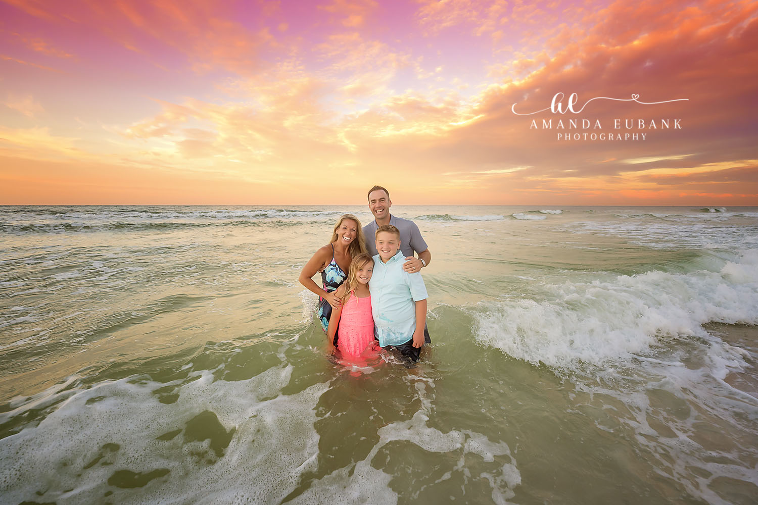 Laughing in the Breaking Waves | Family Photographer in Tampa, Florida Family beach portraits in Tampa, FL showing a family of four crouched laughing as waves break around them.