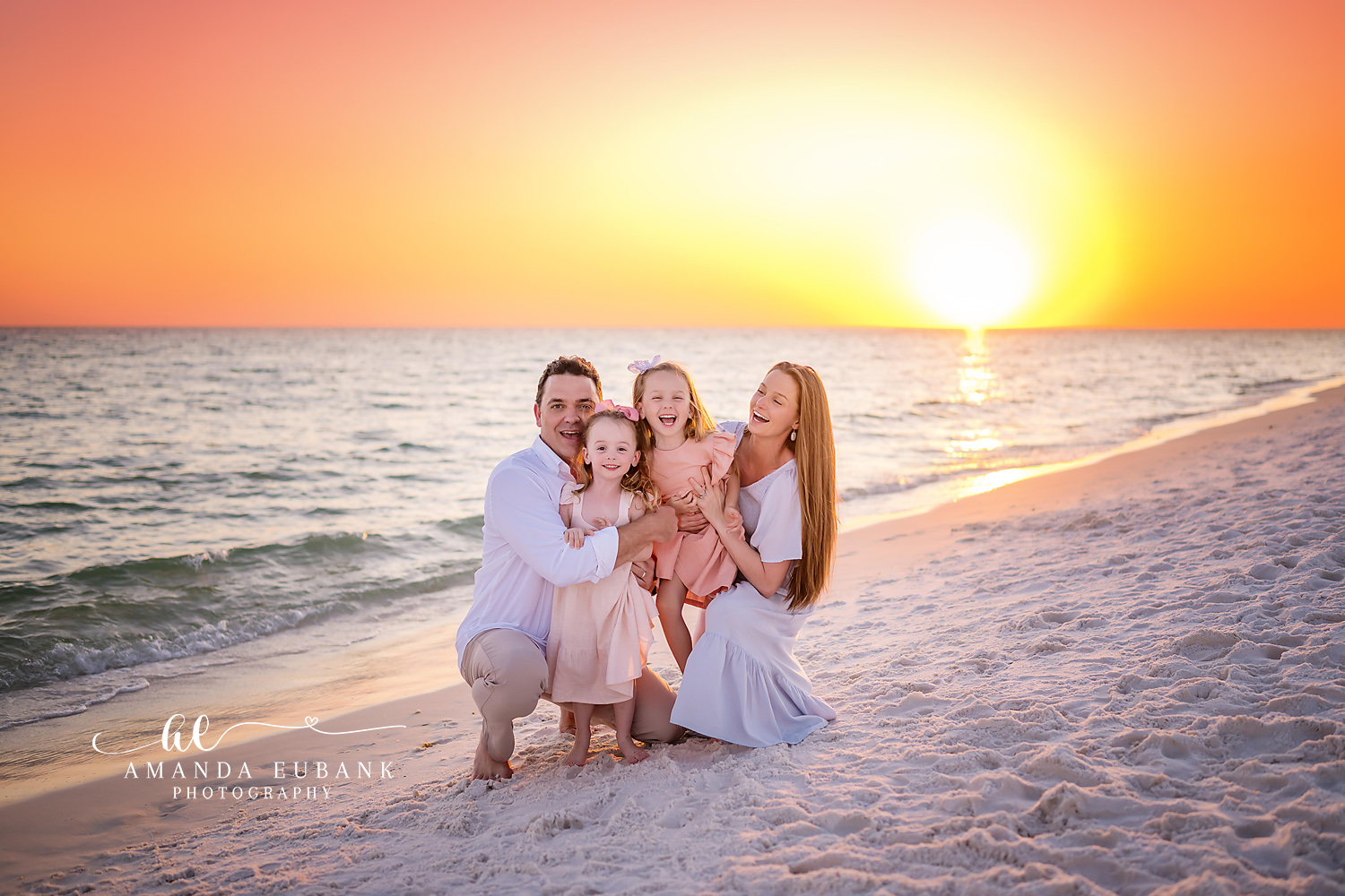 Family Kneeling at Sundown | Tampa Beach Family Photography Tampa family photographer captures a family of four kneeling together on the sand as the sun blazes on the horizon.