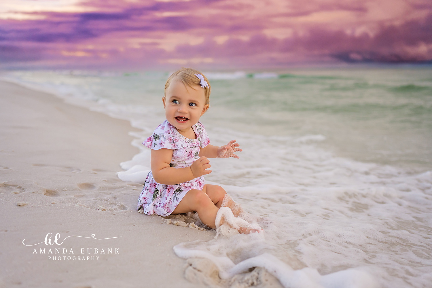 Baby Girl on the Sand | Tampa Baby & Milestone Photographer A baby girl in a floral dress sitting on the sand beside the turquoise water during a baby milestone session in Tampa, Florida.