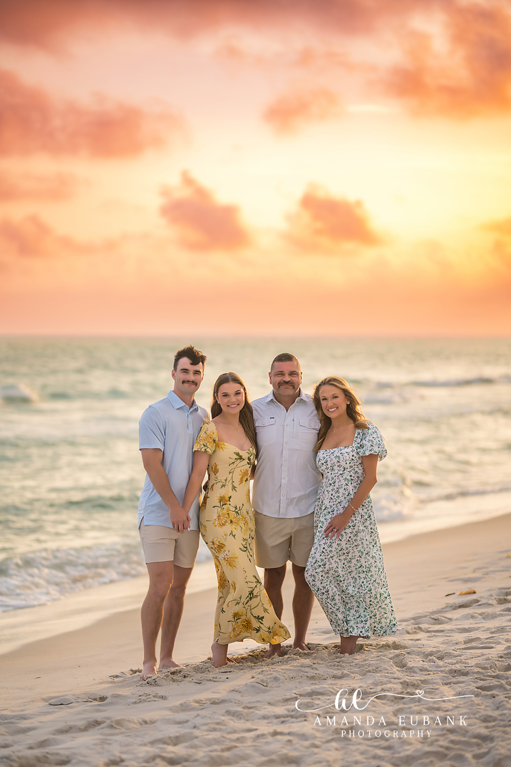 Family of Four in Yellow | Family Photographer in Tampa, Florida Family beach portraits in Tampa, FL showing a family of four standing on the sand in yellow and blue under pastel clouds.