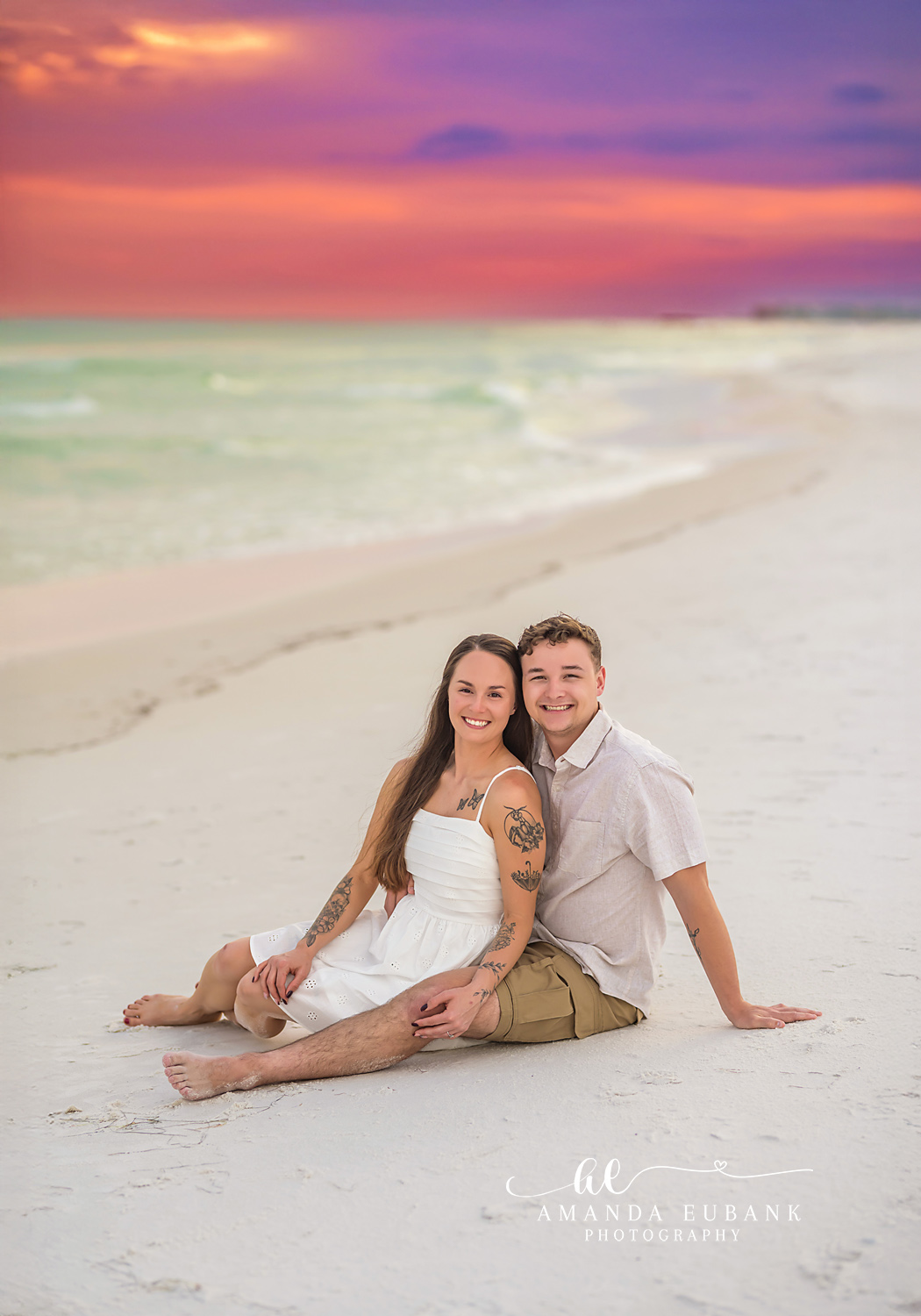 Couple Sitting on the Sand | Tampa Beach Couples Photography Tampa couples photographer captures a couple sitting together on the sand under a coral and purple sky.