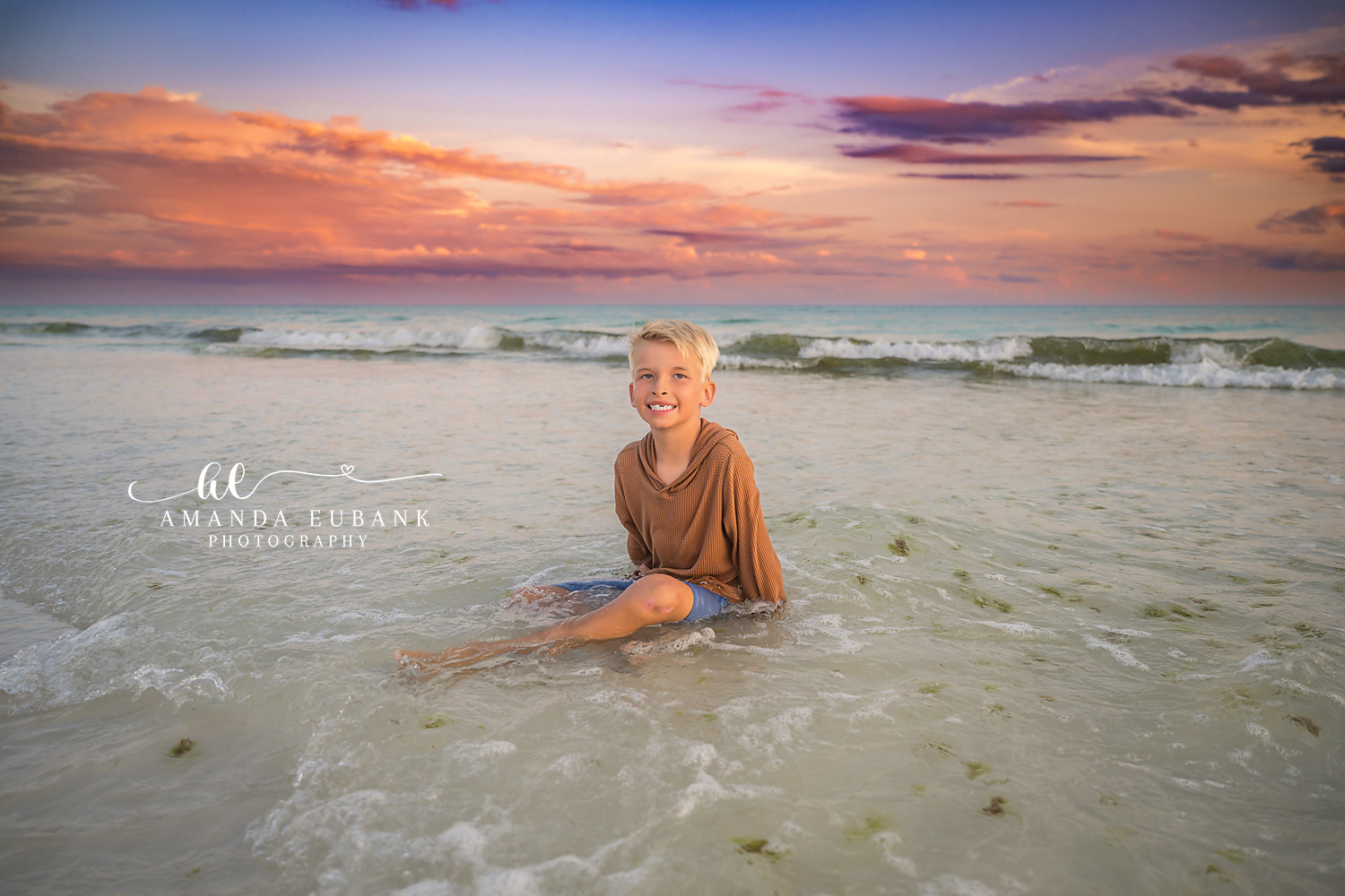 Boy Sitting in the Shallow Water | Tampa Children’s Photographer A young boy sitting in the shallow Gulf water as the sky turns coral at sunset during a children’s portrait session in Tampa, Florida.