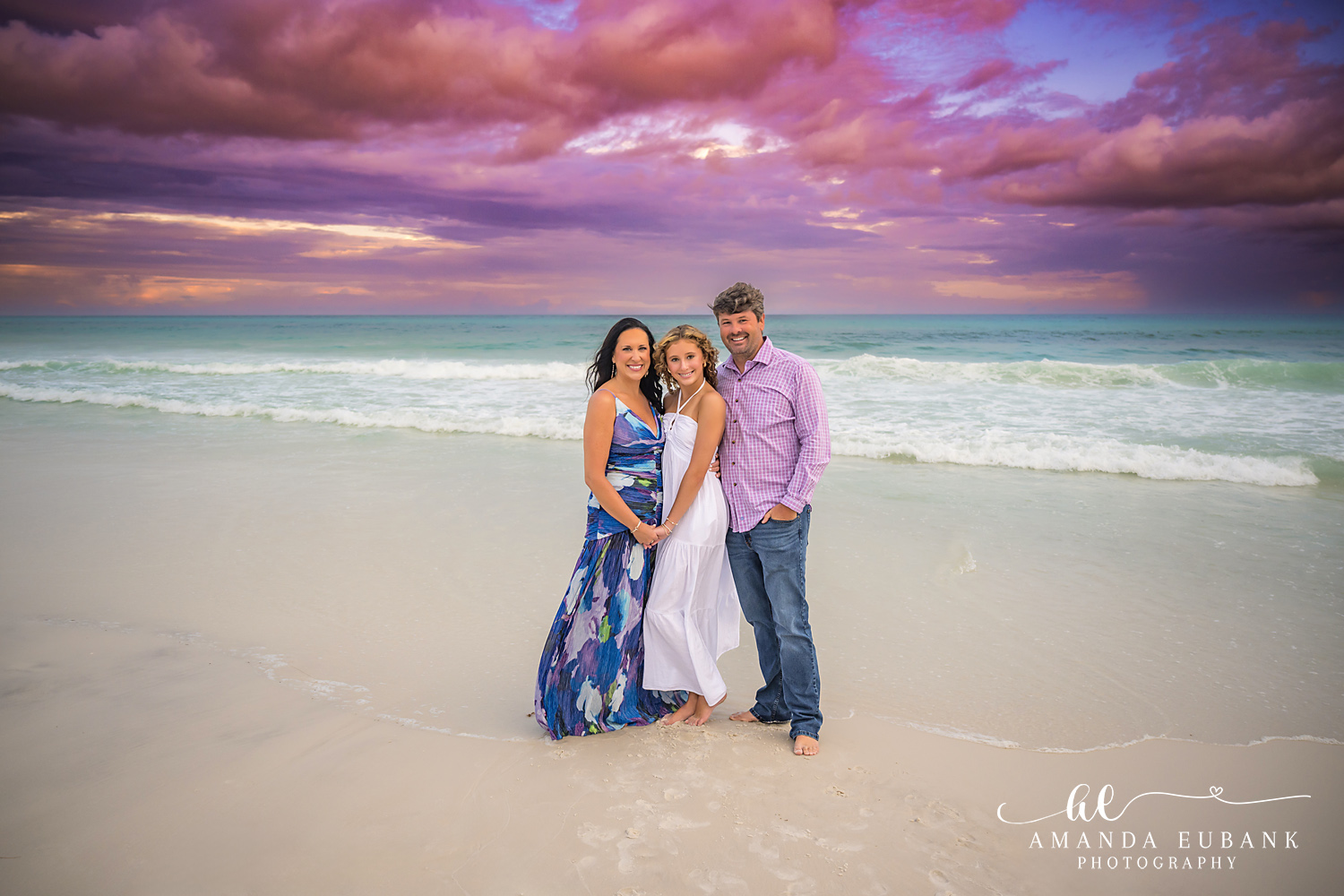 Family of Three at the Shoreline — Tampa Bay Family Portraits A family of three standing on the sand in navy and pink beneath a violet sky — beach family photography in Tampa Bay.