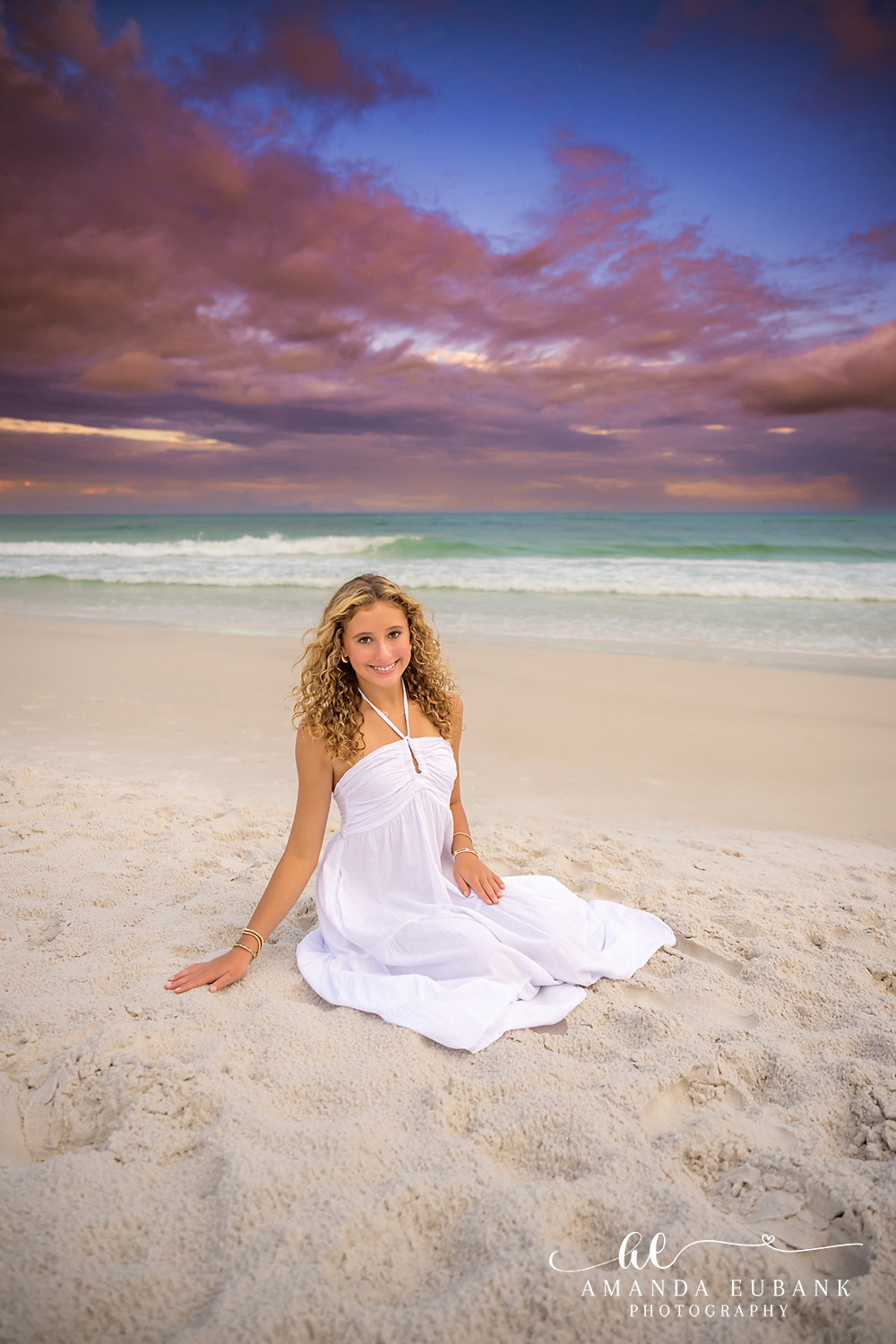 Young Woman in White on the Sand — Tampa Bay Family Portraits A young woman in a white gown sitting on the sand beneath a purple cloudscape — beach family photography in Tampa Bay.