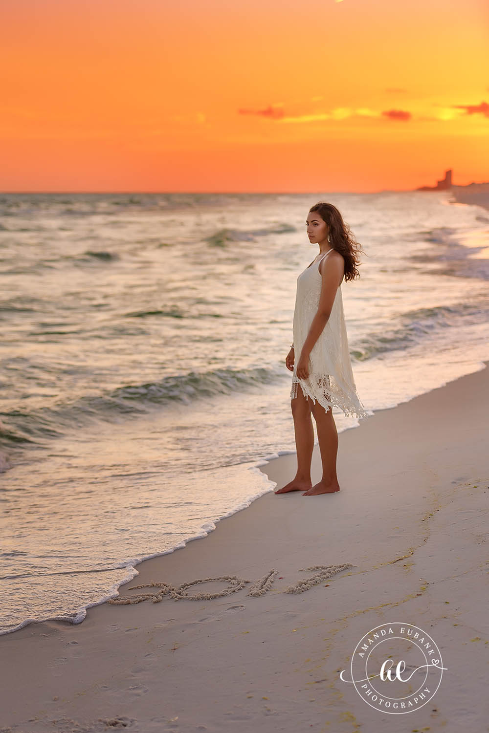 Senior Walking the Shoreline | Beach Senior Session in Tampa, Florida Senior pictures in Tampa Bay showing a senior in a white dress walking the shoreline in warm orange evening light.