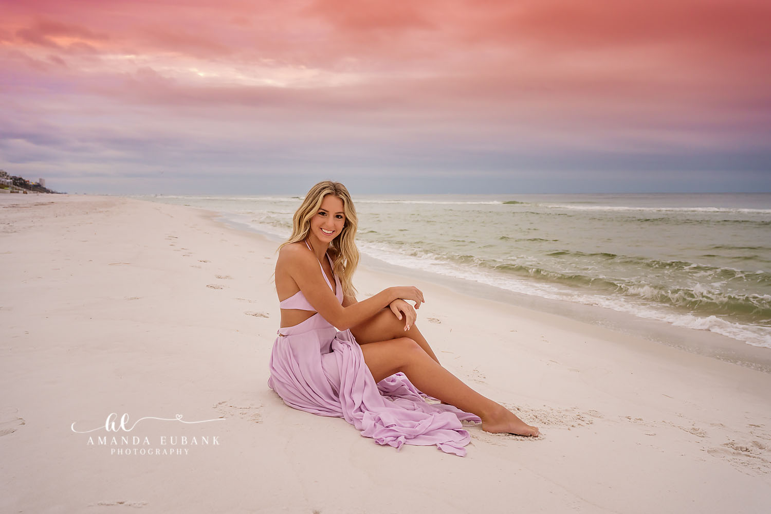 Lavender Skirt on the Sand | Beach Senior Session in Tampa, Florida Senior pictures in Tampa Bay showing a senior in a lavender skirt seated on white sand beneath a pink and blue sky.