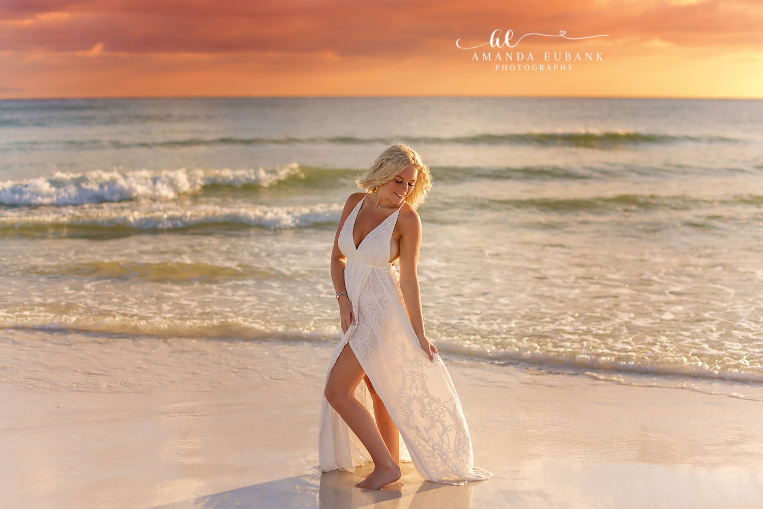 Senior at the Water’s Edge — Tampa Bay Senior Photography A senior in a white dress walking through the surf beneath a coral sunset at a beach senior session in Tampa, FL.