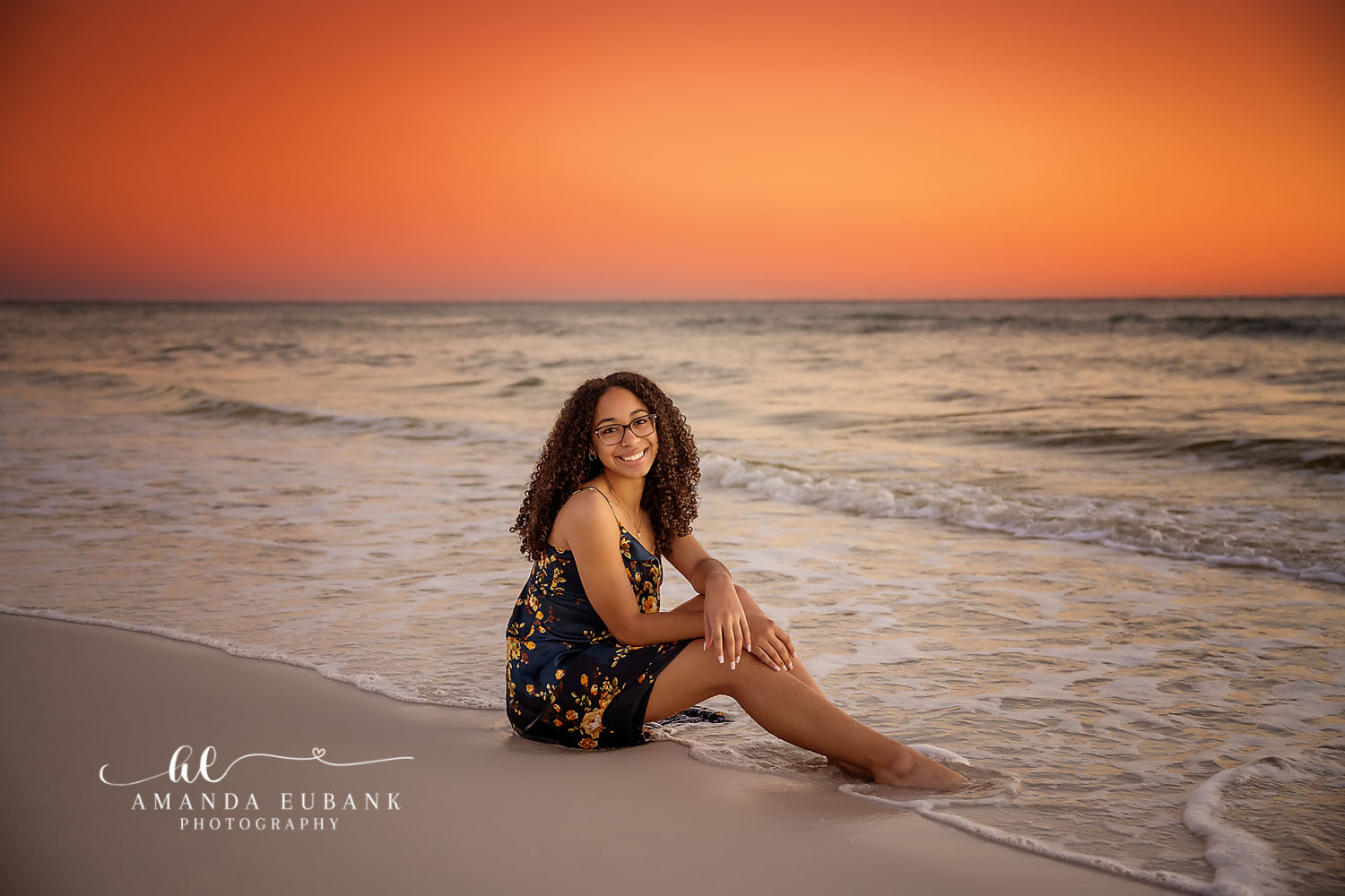 Floral Romper at Sunset | Tampa Senior Photographer A senior in a floral romper seated on the sand under a glowing orange sky during a senior portrait session in Tampa, Florida.