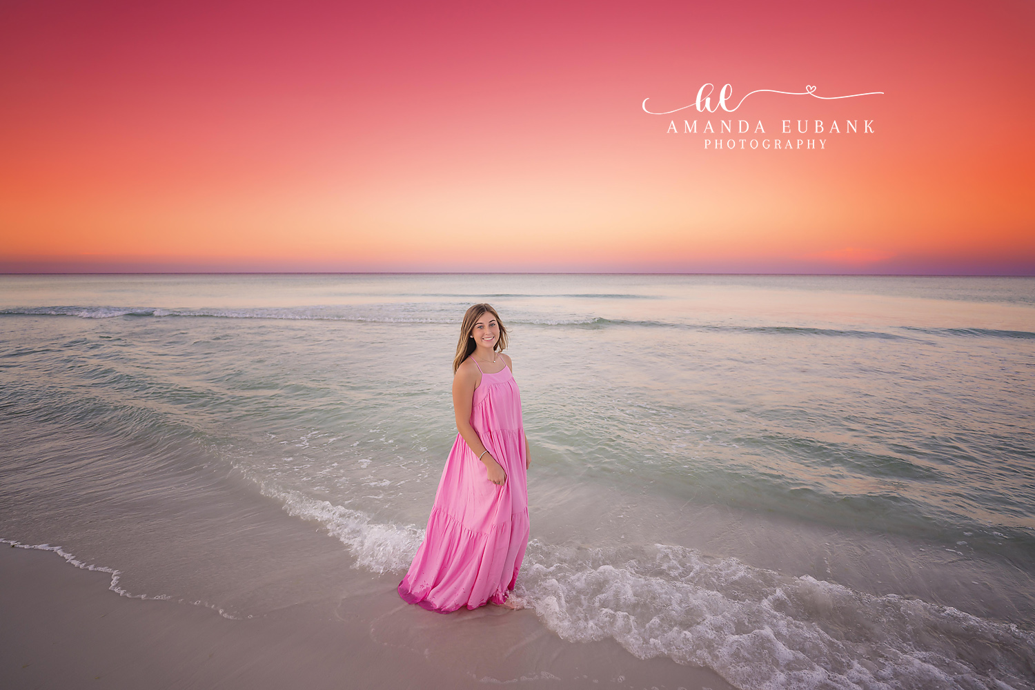 Pink Gown on the Shoreline | Beach Senior Session in Tampa, Florida Senior pictures in Tampa Bay showing a senior in a long pink gown standing on the shoreline beneath a coral sky.