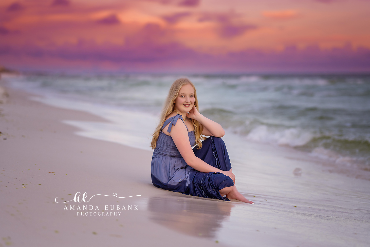 Navy Jumpsuit on the Wet Sand | Tampa Senior Photographer A senior in a navy jumpsuit seated on the wet sand under a violet and coral sky during a senior portrait session in Tampa, Florida.