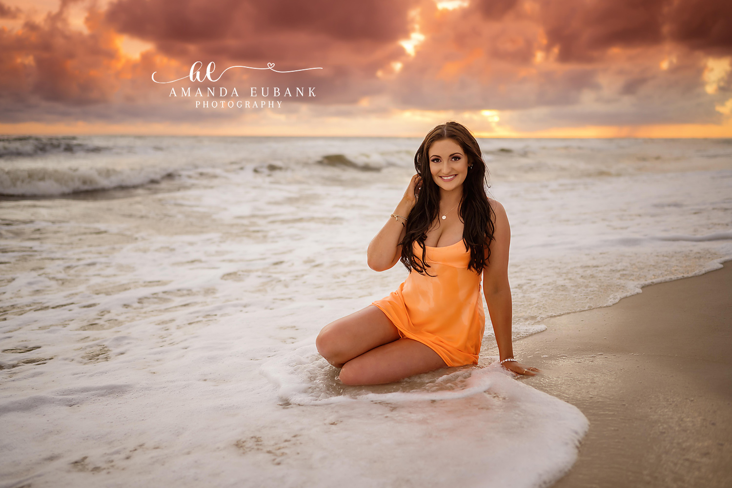 Orange Dress in the Surf — Tampa Bay Senior Photography A senior in an orange dress kneeling in the surf beneath dramatic golden clouds at a beach senior session in Tampa, FL.