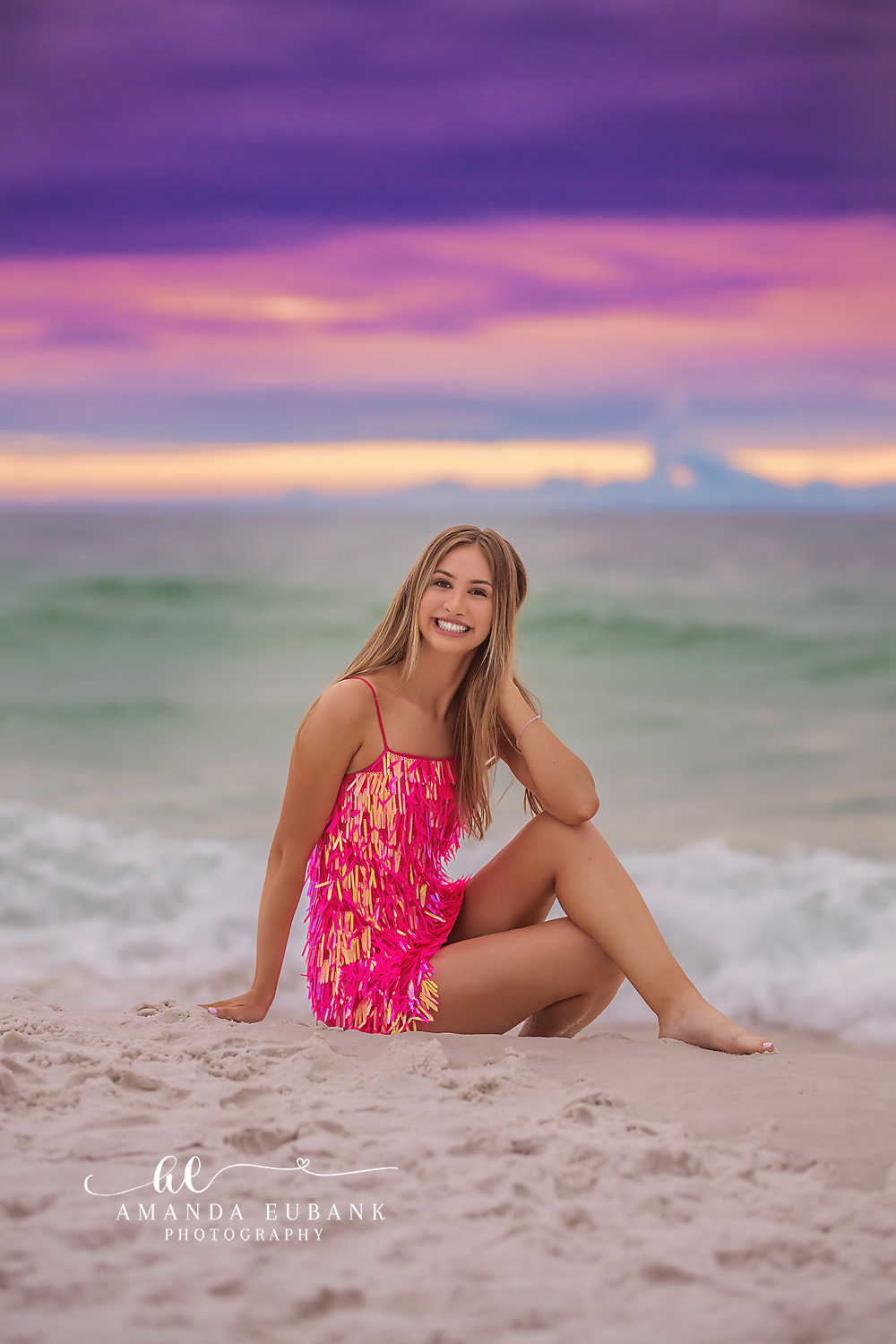 Pink Dress in the Shallows | Beach Senior Session in Tampa, Florida Senior pictures in Tampa Bay showing a senior in a pink patterned dress seated in the shallows under a purple sky.