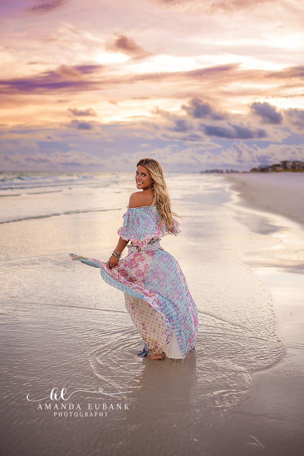 Crouched in the Shallows — Tampa Bay Senior Photography A senior in a floral skirt crouched in the shallow surf beneath pink clouds at a beach senior session in Tampa, FL.