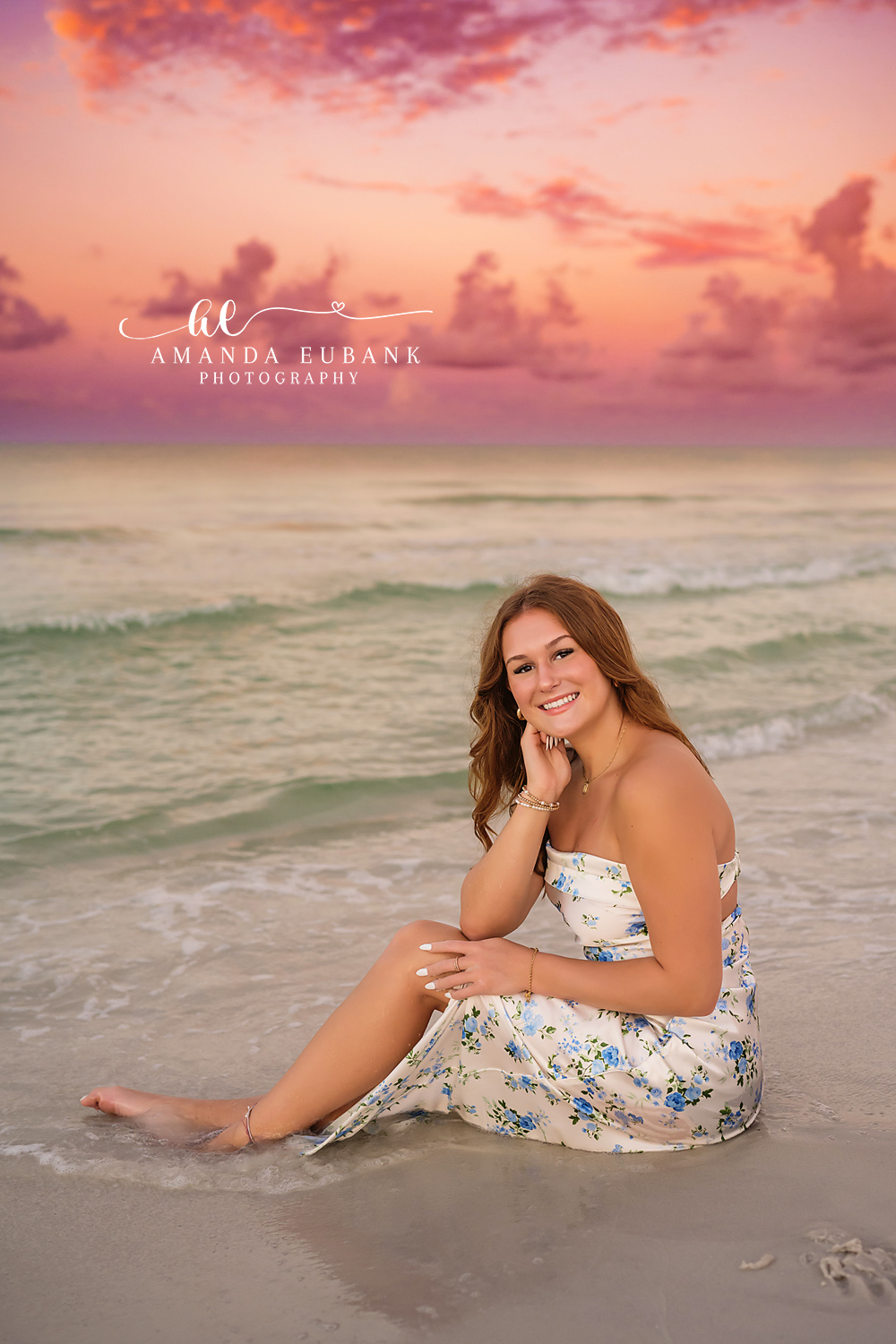 Pink Clouds Senior Portrait — Tampa Bay Senior Photography A senior in a floral dress seated on the sand beneath towering pink clouds at a beach senior session in Tampa, FL.