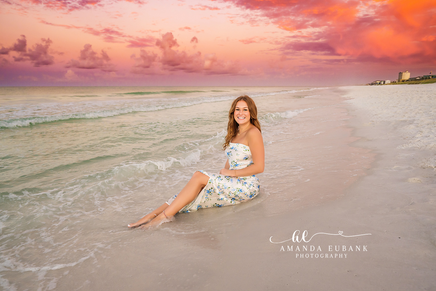 Coral Clouds on the Shoreline | Beach Senior Session in Tampa, Florida Senior pictures in Tampa Bay showing a senior in a floral dress seated on the shoreline under coral sunset clouds.