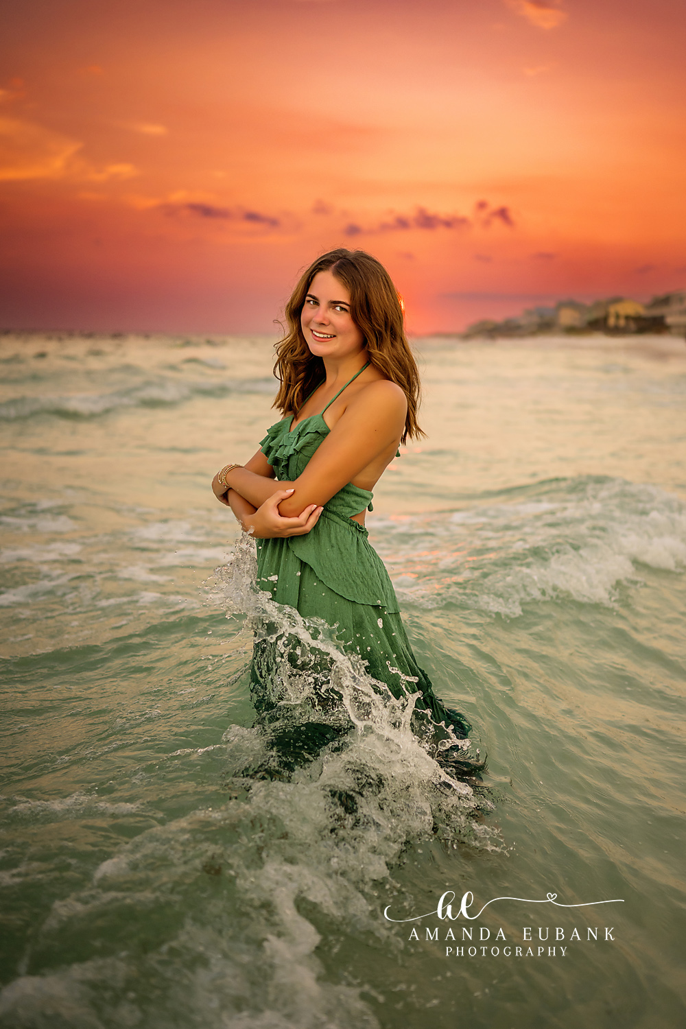 Green Gown in the Waves | Beach Senior Session in Tampa, Florida Senior pictures in Tampa Bay showing a senior in a green gown standing in the waves under a blazing orange sky.