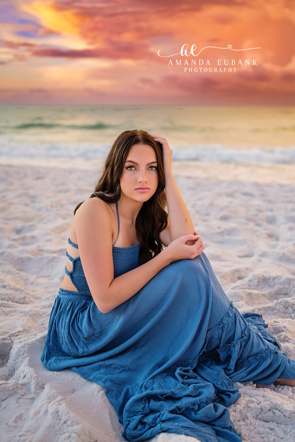 Seated in a Blue Gown — Tampa Bay Senior Photography A senior in a blue gown seated on white sand as the sky glows soft orange at a beach senior session in Tampa, FL.