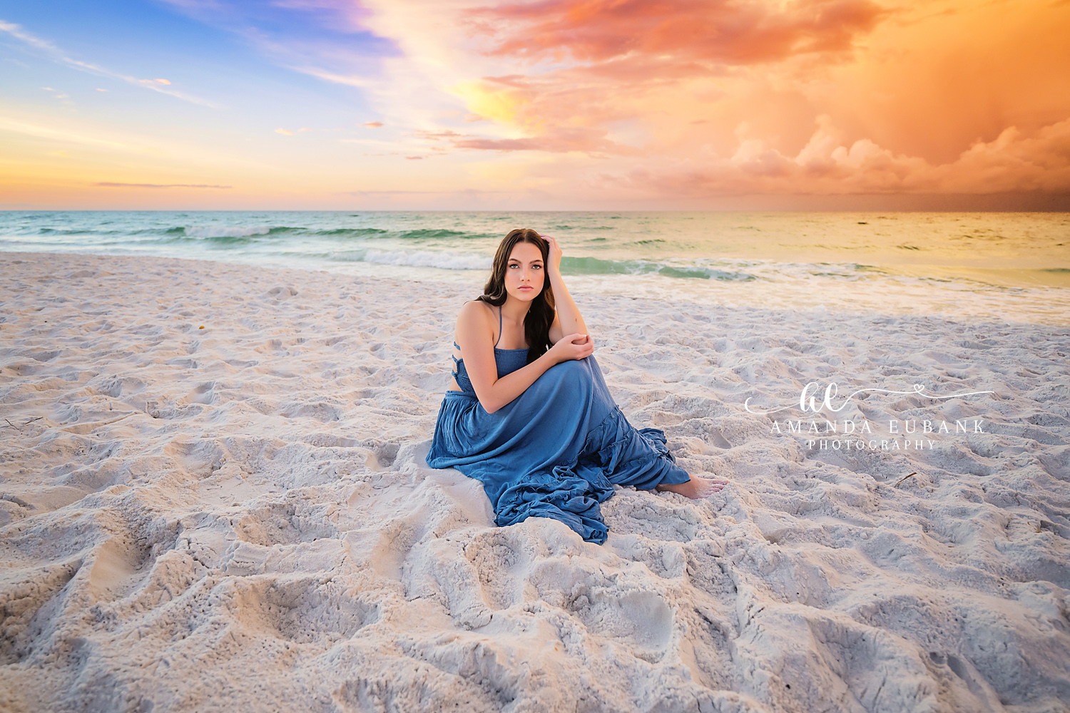 Blue Gown Under a Gold Sky | Beach Senior Session in Tampa, Florida Senior pictures in Tampa Bay showing a senior in a blue gown seated on the sand beneath streaks of gold cloud.