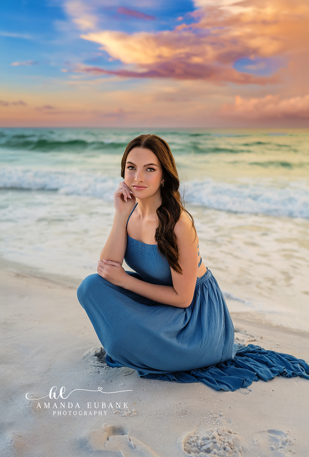 Kneeling in a Blue Gown | Tampa Senior Photographer A senior kneeling on the sand in a blue gown beneath soft pastel light during a senior portrait session in Tampa, Florida.