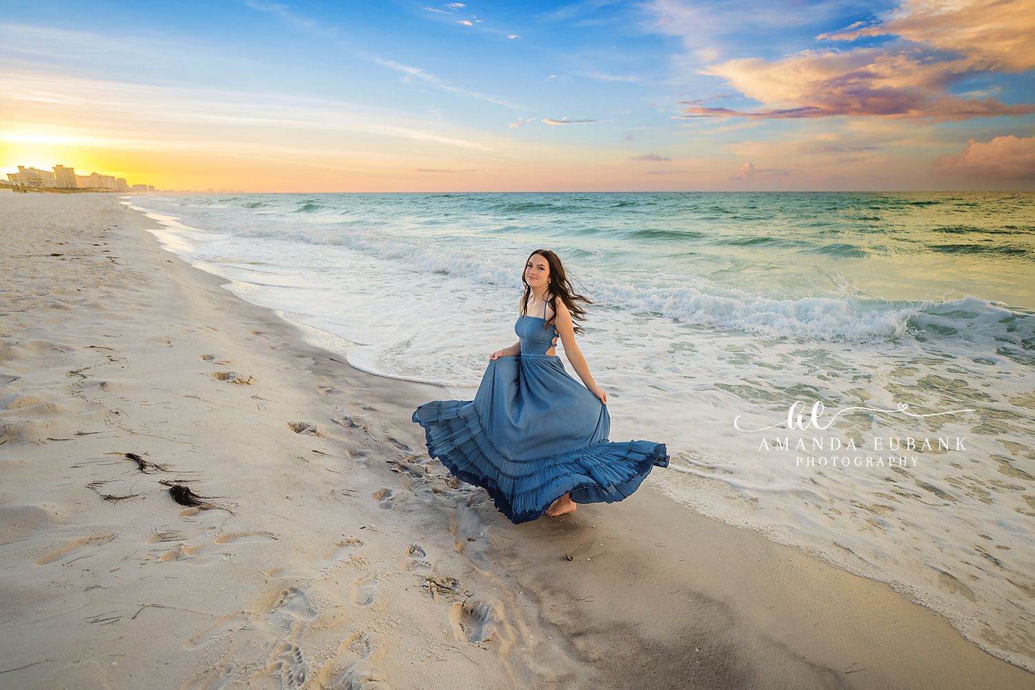 Walking the Sand at Sundown | Beach Senior Session in Tampa, Florida Senior pictures in Tampa Bay showing a senior in a blue gown walking the sand as the sun drops toward the water.