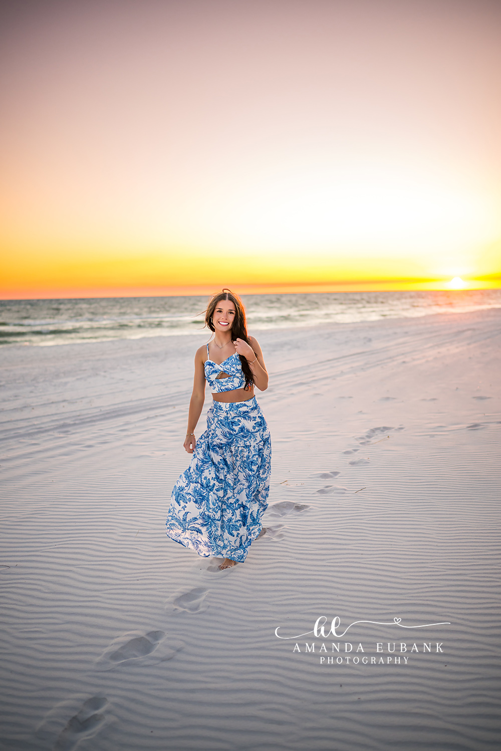 Walking Toward the Sun — Tampa Bay Senior Photography A senior in a blue floral skirt walking the sand as the sun sits on the water at a beach senior session in Tampa, FL.