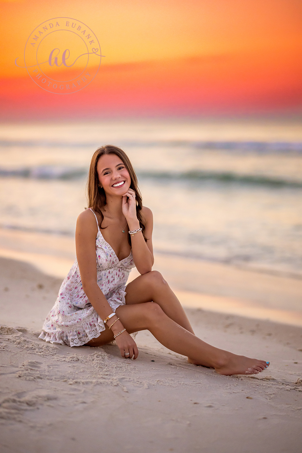 Seated Under a Red Sky | Beach Senior Session in Tampa, Florida Senior pictures in Tampa Bay showing a senior in a white lace dress seated on the sand beneath a red and orange sky.