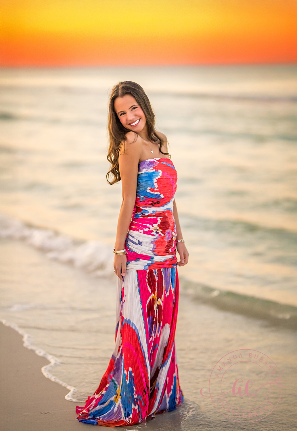 Pink Gown at the Water’s Edge | Tampa Senior Photographer A senior in a pink patterned gown standing at the water's edge at sunset during a senior portrait session in Tampa, Florida.