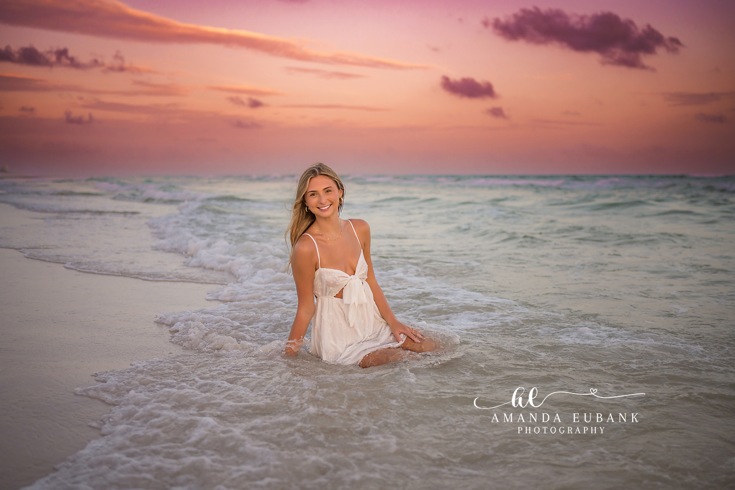 Seated in the Rolling Surf | Beach Senior Session in Tampa, Florida Senior pictures in Tampa Bay showing a senior in a white dress seated in the rolling surf under a pink sky.