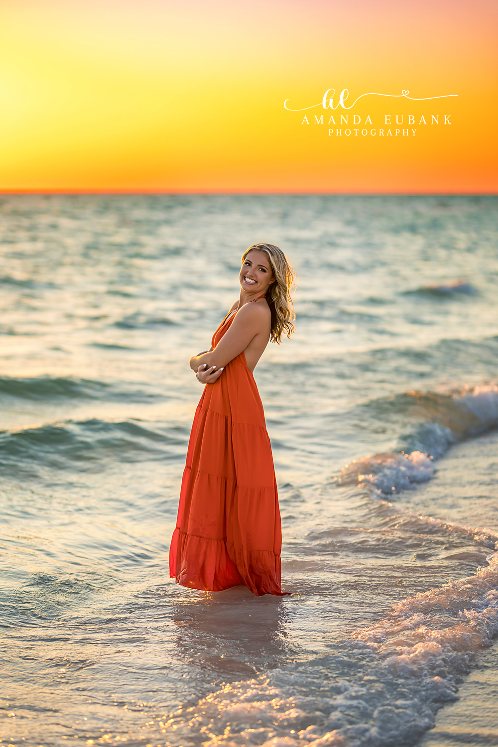 Red Gown in the Shallows | Beach Senior Session in Tampa, Florida Senior pictures in Tampa Bay showing a senior in a red gown standing in the shallow water under a golden sky.