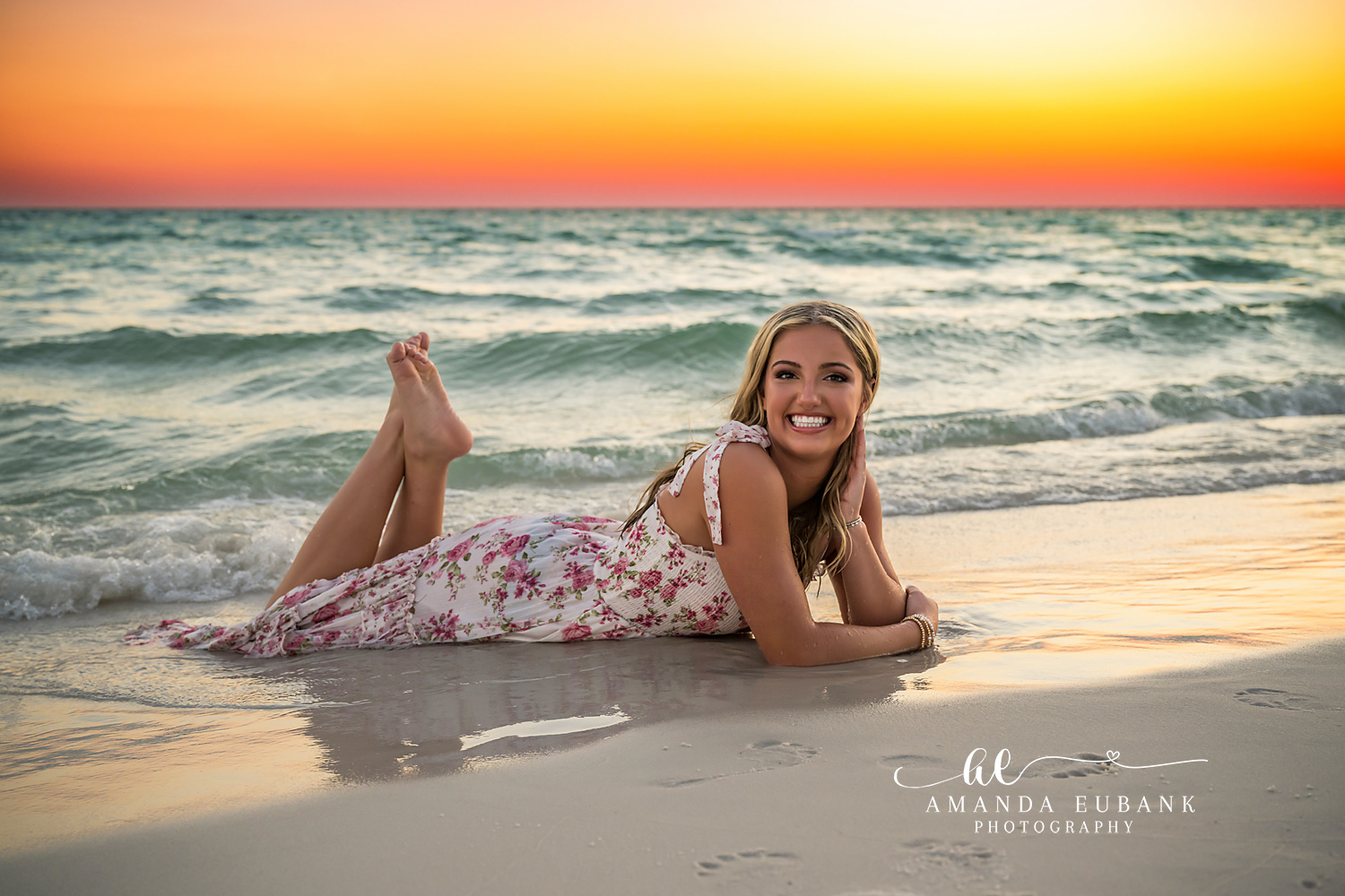 Smiling on the Shoreline | Tampa Senior Photographer A smiling senior in a floral dress lying on the shoreline at sunset during a senior portrait session in Tampa, Florida.