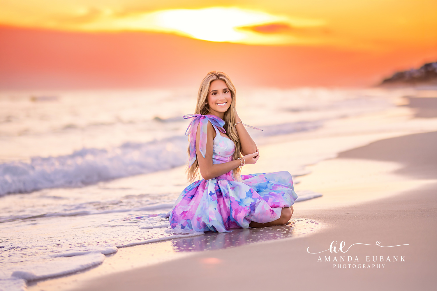Seated in the Orange Glow | Beach Senior Session in Tampa, Florida Senior pictures in Tampa Bay showing a senior in a printed gown seated on the sand under a fiery orange sky.