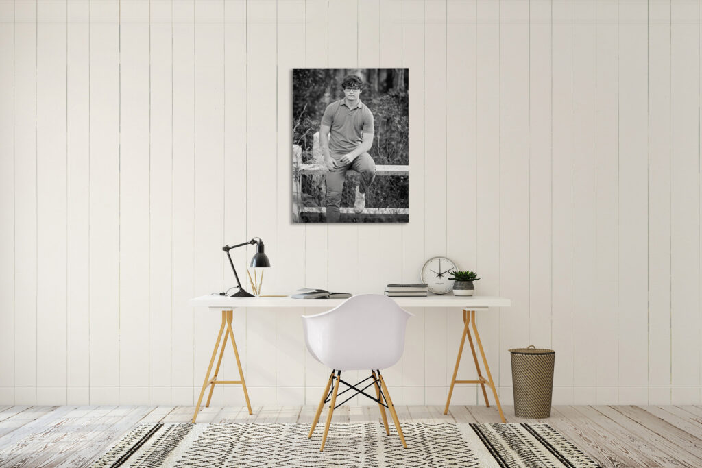 Black and white canvas of a senior guy seated on a wooden fence, displayed above a white desk